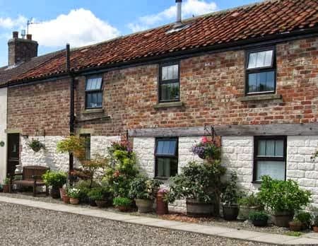 Charming stone cottage with a red-tiled roof, surrounded by potted plants and a gravel path in a serene setting.