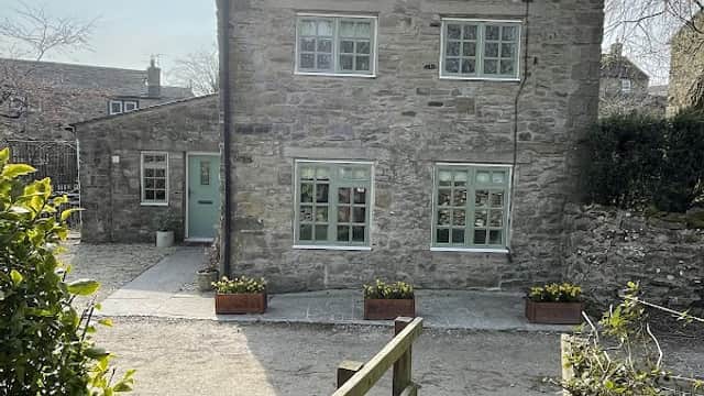 Stone cottage with light blue windows and flower boxes, surrounded by greenery and a wooden walkway.