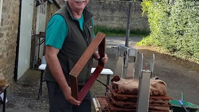 A smiling man holds a wooden frame near tables with trays and buckets in a farmyard setting surrounded by greenery.