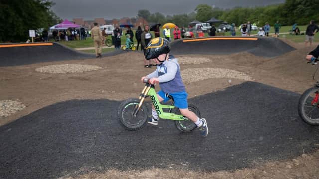 A child on a green balance bike rides over a pump track, with spectators and tents in the background.