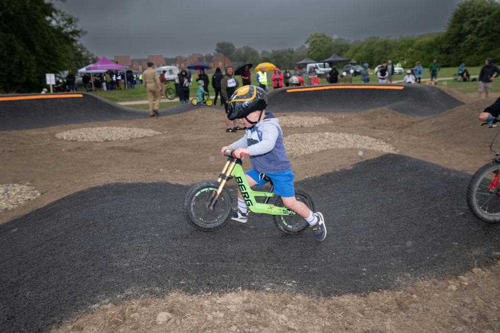 A child on a green balance bike rides over a pump track, with spectators and tents in the background.