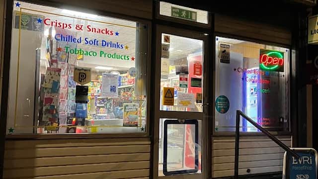 Newsagent storefront with illuminated signs, displaying snacks, drinks, and tobacco products, in a corner shop setting.