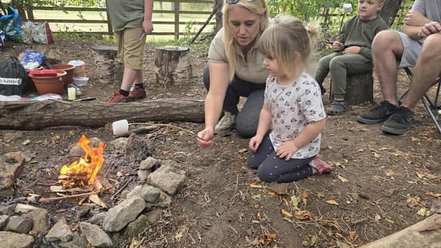 A woman teaches a young girl by a small campfire, surrounded by children and nature in a woodland setting.