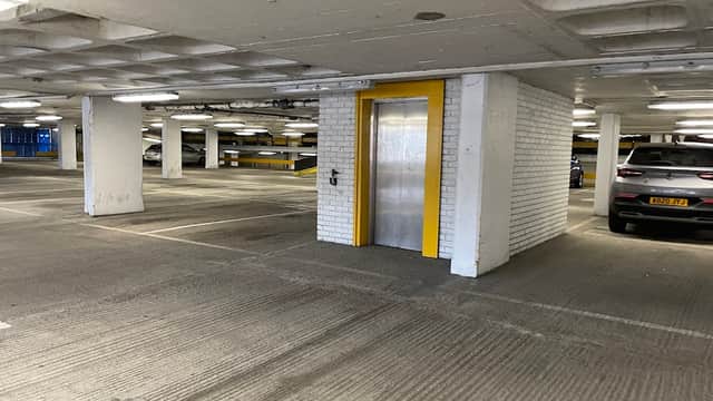 Elevator entrance with yellow door in a spacious car park, parked cars visible in the background.