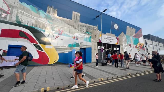 Crowd of people walking past a colorful mural and entrance to York Community Stadium, with parked cars nearby.