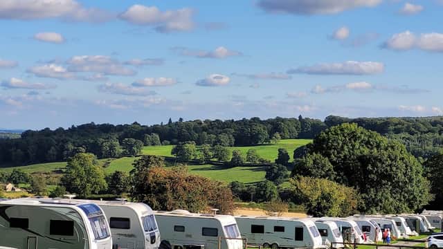 Caravans lined up on a grassy area with rolling hills and trees in the background under a blue sky with fluffy clouds.