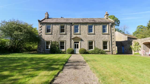 Two-story stone manor with chimneys, surrounded by lush green lawn, gravel path leading to entrance, and blue sky.