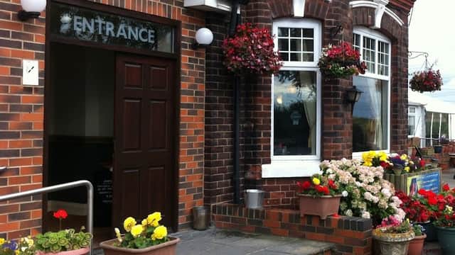 Brick building entrance with a sign, surrounded by colorful flower pots and hanging baskets.