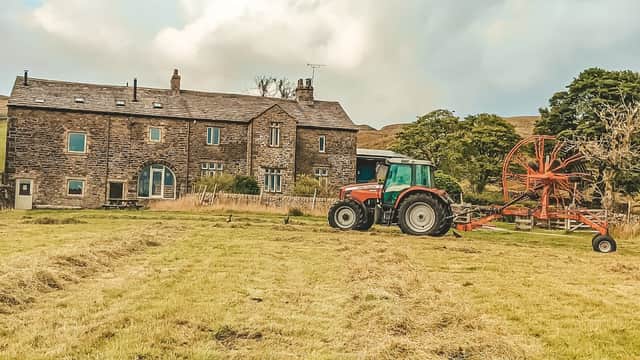 Stone farmhouse with large windows, surrounded by fields, and a red tractor with a hay rake in the foreground.