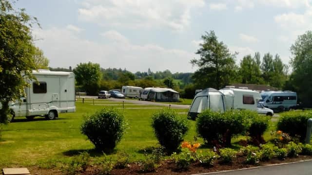 Caravans parked on a grassy area, surrounded by trees and flower beds under a partly cloudy sky.