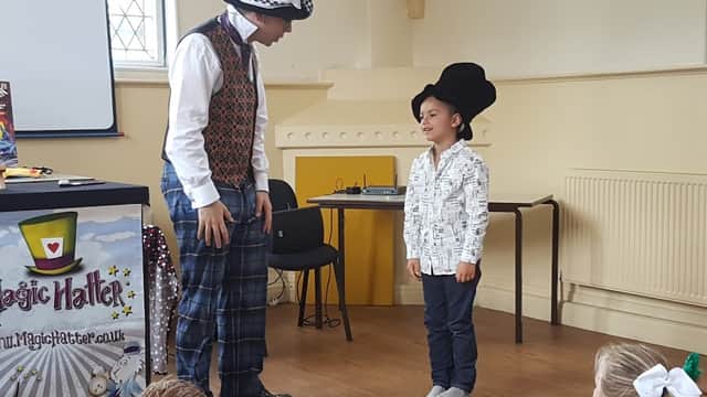 A magician performs for a child wearing a top hat, with an audience of children seated in the background.