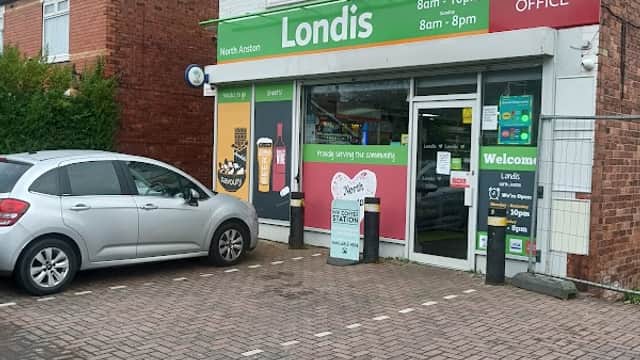 Londis storefront with a Post Office sign, open hours displayed, and a gray car parked in front on a brick pavement.