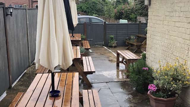 Wooden picnic tables with umbrellas in a wet outdoor area, surrounded by greenery and a low fence.