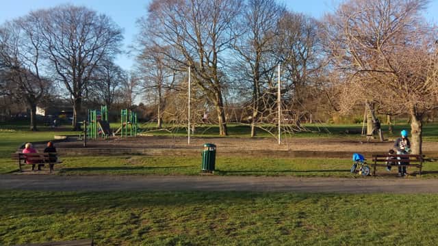 Wellholme Park features playground equipment, benches, trees, and a grassy area under a clear blue sky.