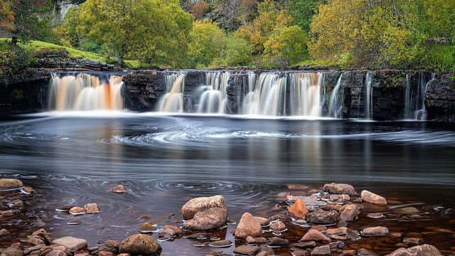 Waterfall cascading over rocks, surrounded by vibrant autumn foliage and a serene river in West Stonesdale, Yorkshire.