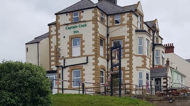 Victorian-style building with green signage reading "Captain Cook Inn," surrounded by grass and a cloudy sky.