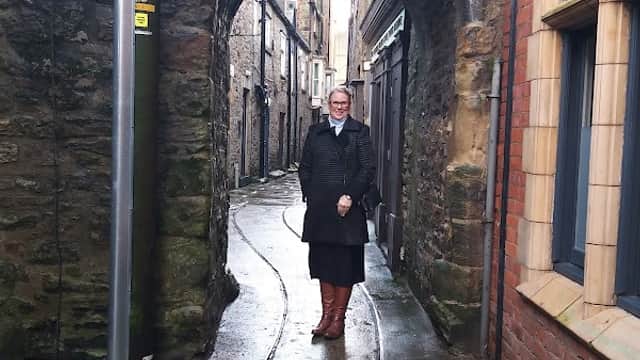Narrow stone alleyway with a person standing under an arch, flanked by brick and stone buildings, wet pavement.