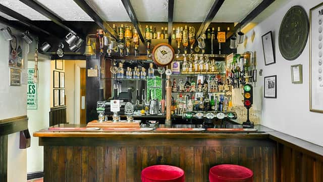 Wooden bar with red stools, shelves lined with bottles, and a clock on the wall in a cozy pub setting.
