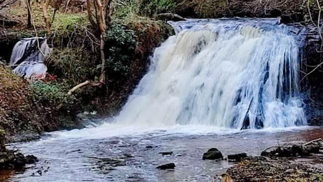 Waterfall cascading over rocks, surrounded by trees and greenery, with a flowing river in the foreground.