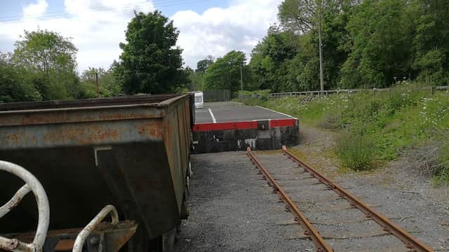 Rusty train car on tracks with a platform in the background, surrounded by greenery and trees under a cloudy sky.