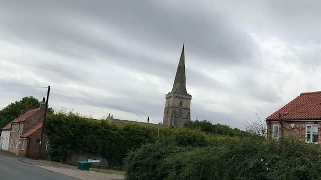 Village hall with a church spire in the background, surrounded by greenery and a quiet road in Ottringham, Yorkshire.