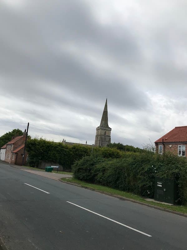 Village hall with a church spire in the background, surrounded by greenery and a quiet road in Ottringham, Yorkshire.