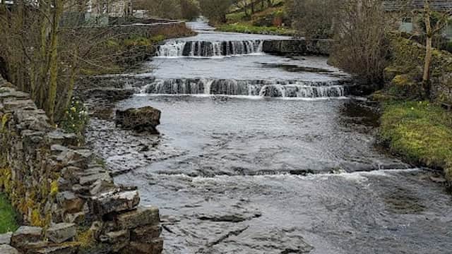 A serene river flows past a stone wall, with a small waterfall and trees lining the banks under a cloudy sky.