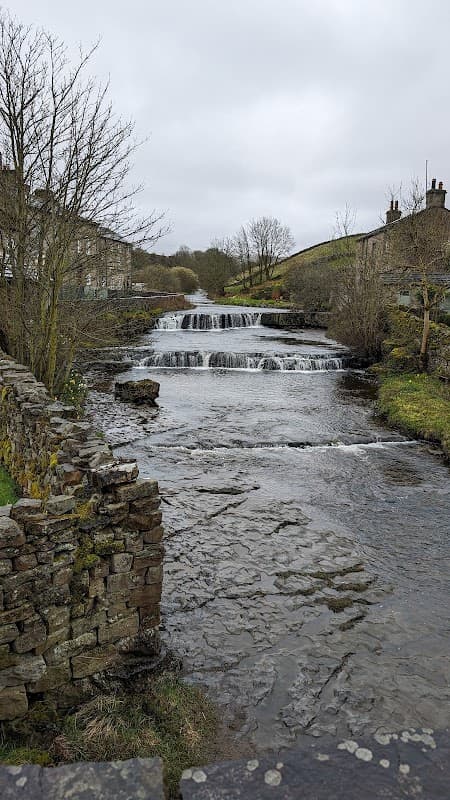 A serene river flows past a stone wall, with a small waterfall and trees lining the banks under a cloudy sky.