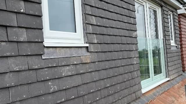 Newly installed windows on a grey shingled wall, with a roof edge visible below and cloudy sky above.