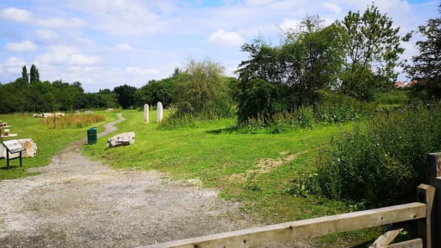 Lush green landscape with a gravel path, stone markers, and wooden gate under a blue sky with fluffy clouds.