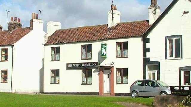 The White Horse Inn with a green sign, white walls, and a car parked beside a pond under a blue sky.