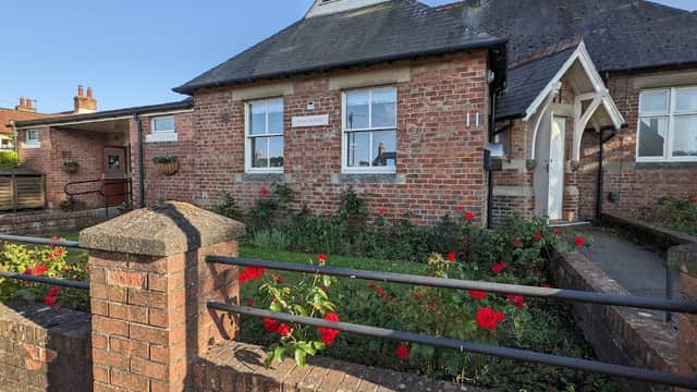 Brick building with a gabled roof, white trim, and a garden of red roses in front. Clear blue sky above.