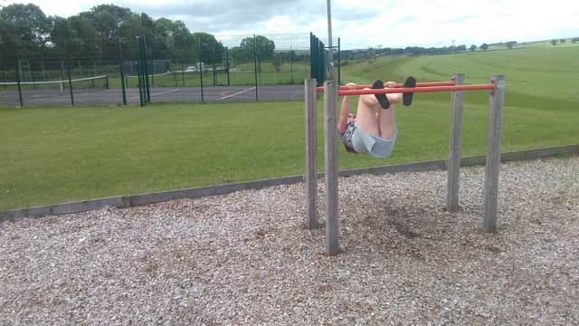 A child hangs upside down on a horizontal bar in a grassy field with a tennis court in the background.