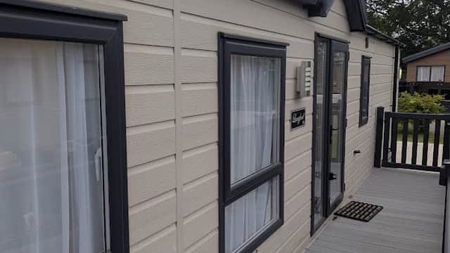 Exterior view of a holiday park cabin with gray siding, windows, and a wooden deck in a green, tree-lined setting.