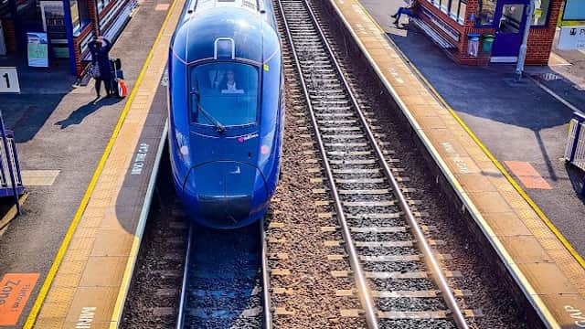 A blue train at Brough station, with platforms, tracks, and passengers waiting nearby under clear skies.