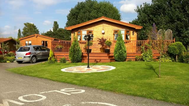 Wooden holiday cabin with a deck, surrounded by greenery and decorative plants, on a well-maintained grassy area.