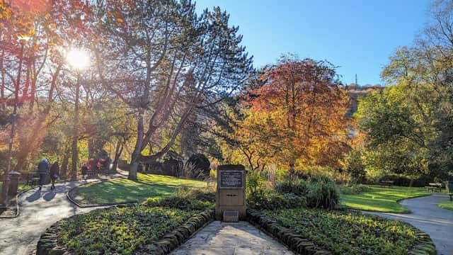 A serene memorial garden with vibrant autumn foliage, stone pathways, and a central plaque under a bright blue sky.