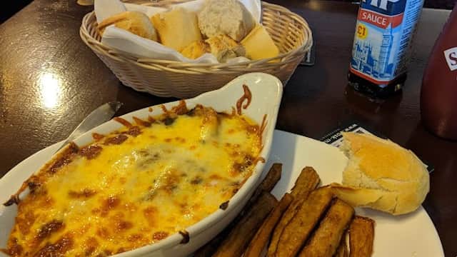 A plate of cheesy lasagna with fries, a roll, and a basket of bread beside a bottle of HP sauce on a wooden table.