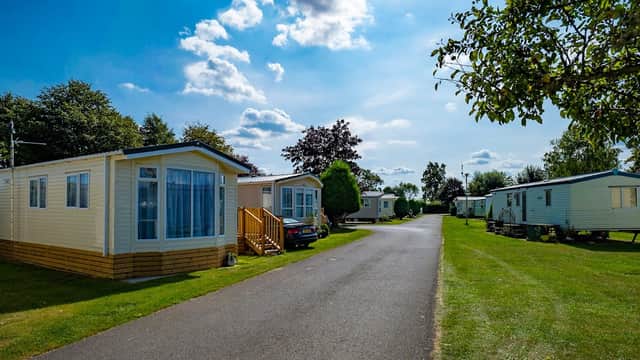 Caravans lined along a paved path in a green holiday park under a blue sky with scattered clouds.