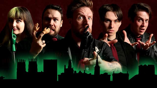 Five performers in dark attire pose dramatically against a shadowy city skyline, evoking a spooky atmosphere.