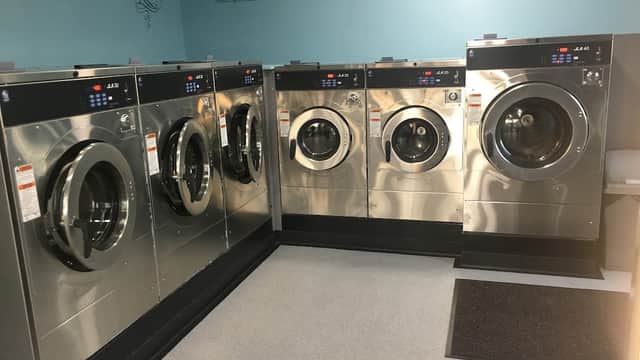 Six modern washing machines in a bright laundry facility with blue walls and pricing signs above.