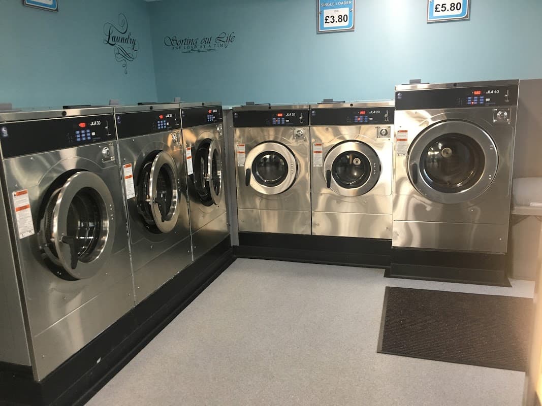 Six modern washing machines in a bright laundry facility with blue walls and pricing signs above.