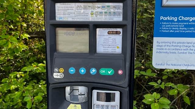 Parking meter with payment options, surrounded by greenery and a sign detailing parking charges and terms.