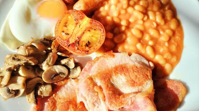 Full English breakfast featuring eggs, sausages, bacon, baked beans, mushrooms, and grilled tomatoes on a plate.