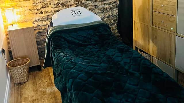 A cozy treatment room featuring a green blanket-covered massage table, wooden storage, and exposed stone walls.