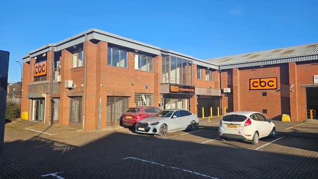 Brick building with large windows and "cbc" signage, surrounded by parked cars in a paved lot under a clear blue sky.
