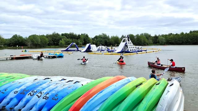 Colorful kayaks stacked by the shore, with people kayaking and inflatable water obstacles in a lake surrounded by trees.