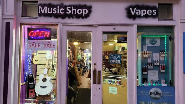 Music shop storefront with guitars, an "OPEN" sign, and vape products displayed in the window.