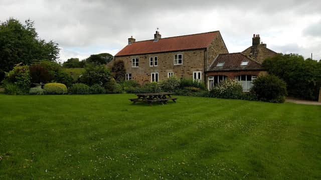 Charming stone hotel with a red-tiled roof, surrounded by lush green gardens and picnic tables under a cloudy sky.