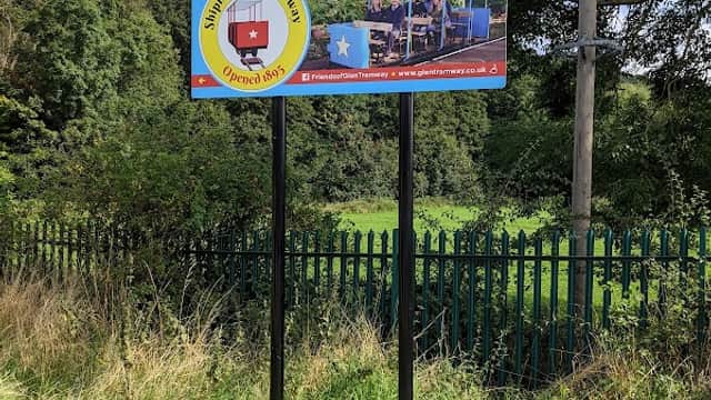 Sign for Shipley Glen Tramway with images and directions, surrounded by greenery and a fence.